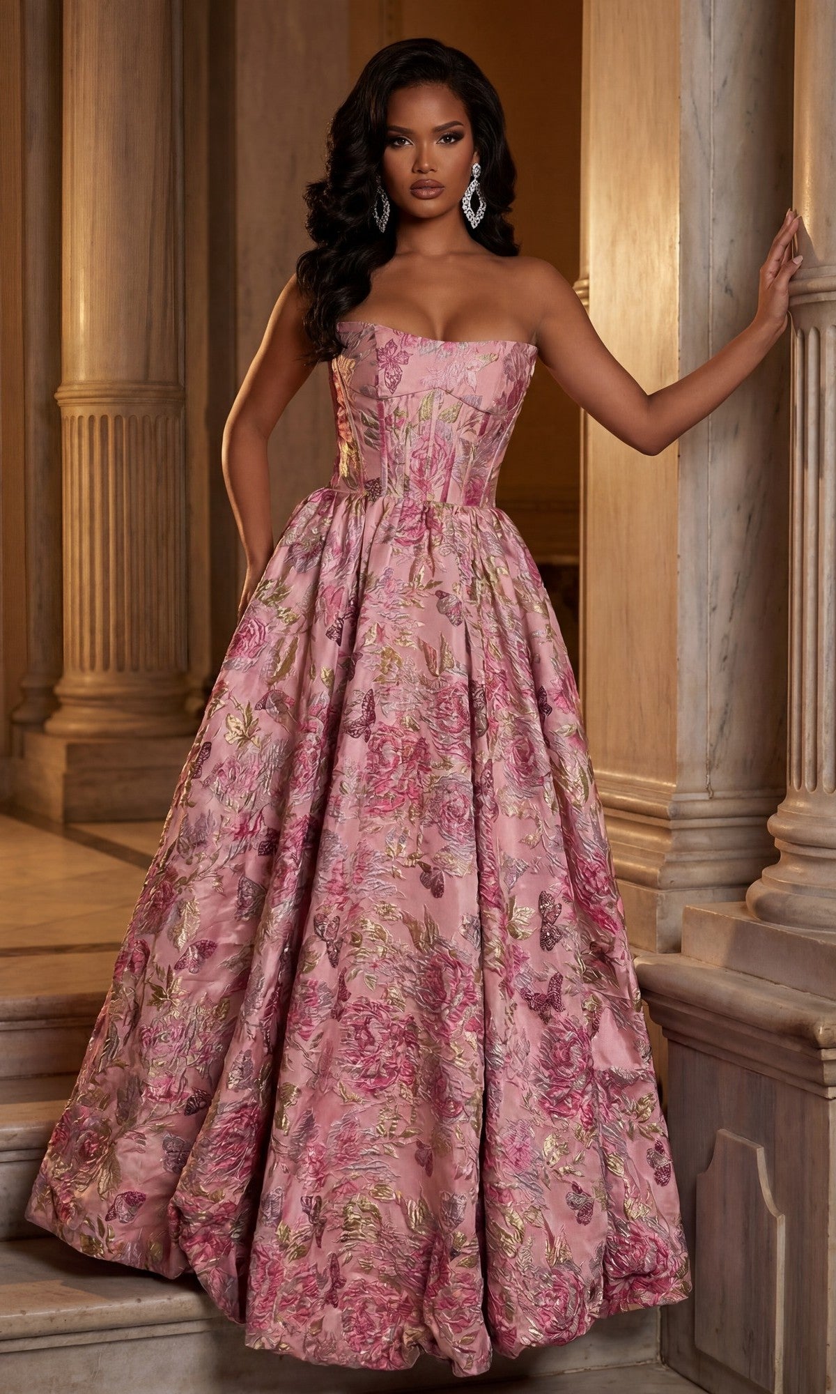 A woman stands by marble columns wearing the Portia and Scarlett XO180 long formal dress in fuchsia/gold, featuring a strapless design, floor-length floral pattern with gold accents, and an elegant A-line skirt.